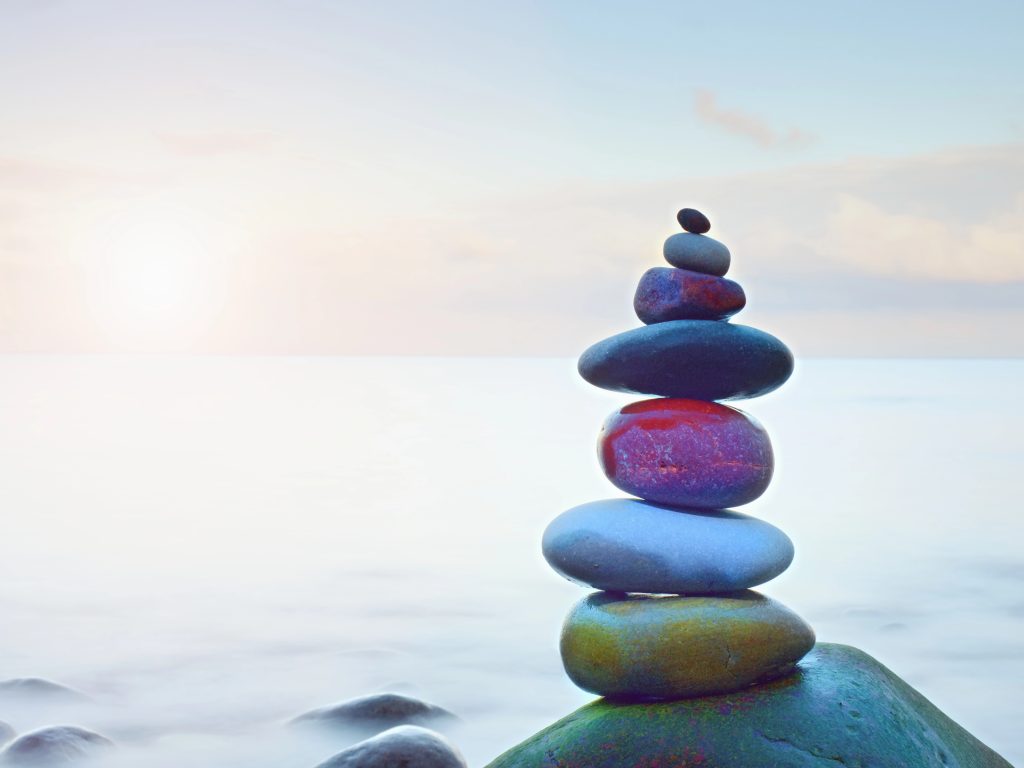 depositphotos 160459246 xl A balanced stack of smooth stones sits on a rock by calm water, the sun low on the horizon and a pastel sky behind—evoking meditation bliss and a peaceful moment for mental health.